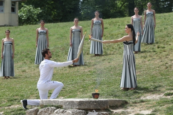 扮演最高女祭司的演員瑪麗·米娜（前右）點(diǎn)燃第一棒火炬手——希臘運(yùn)動員、東京奧運(yùn)會男子賽艇項(xiàng)目金牌得主斯特凡諾斯·恩圖斯科斯手中的火炬。新華社記者 趙丁喆 攝